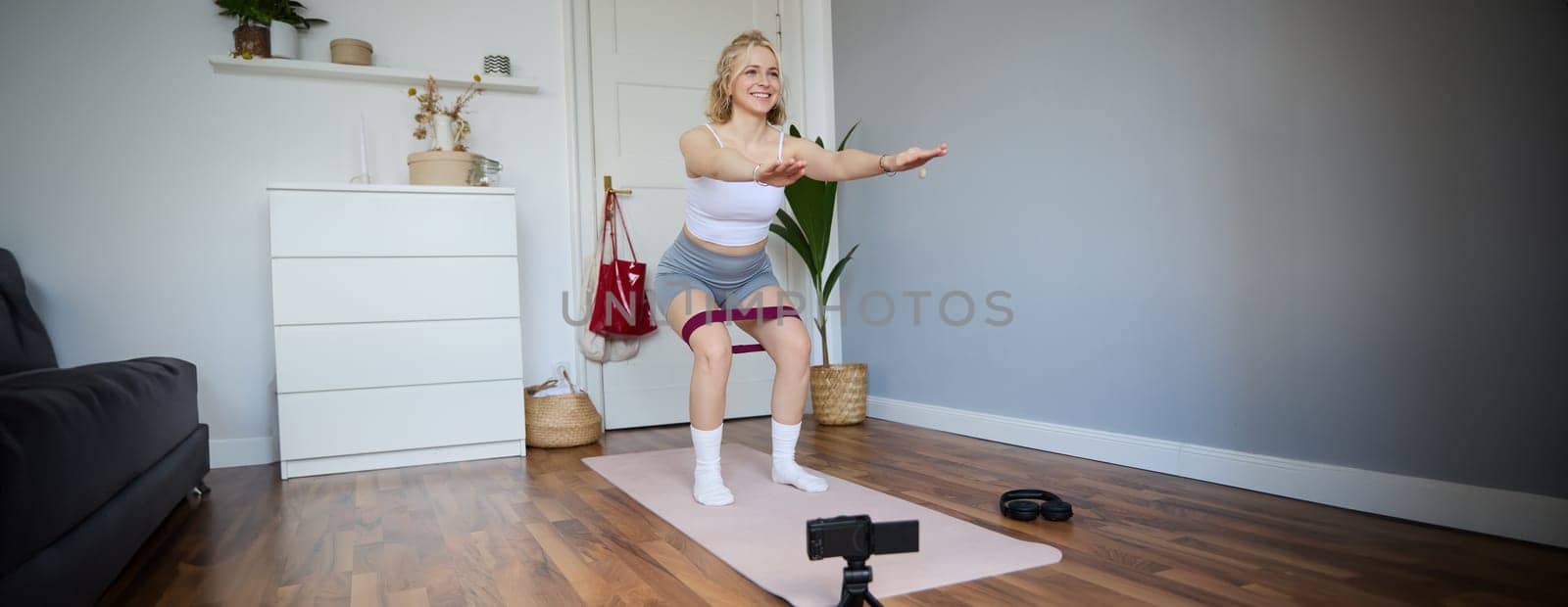 Portrait of young athletic woman, vlogger making a video about home workout, fitness instructor shooting how to do leg exercises, using elastic resistance band, using yoga mat by Benzoix