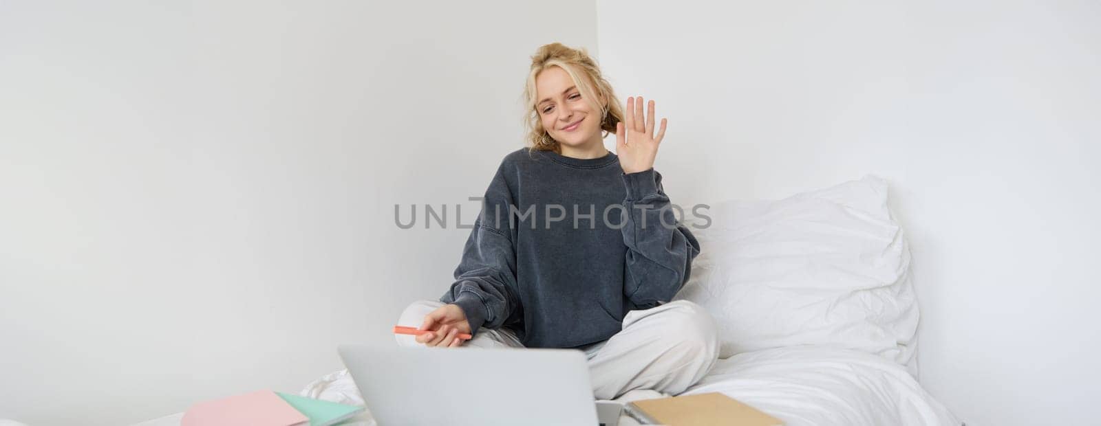 Portrait of beautiful, smiling young woman sitting on bed, waves her hand at laptop camera, says hello to friend on video chat app, studies at home on remote, connects to online course by Benzoix