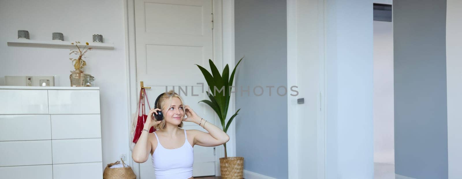 Vertical shot of smiling young woman using video tutorials for workout at home, sitting with laptop in wireless headphones on yoga mat, follows fitness instructor on social media, repeats exercises by Benzoix