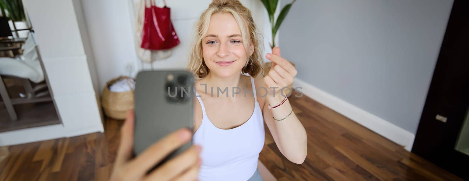 Portrait of young beautiful blond woman, fitness instructor doing exercises at home on yoga mat, taking selfies on smartphone, recording video of herself for social media account about workout.
