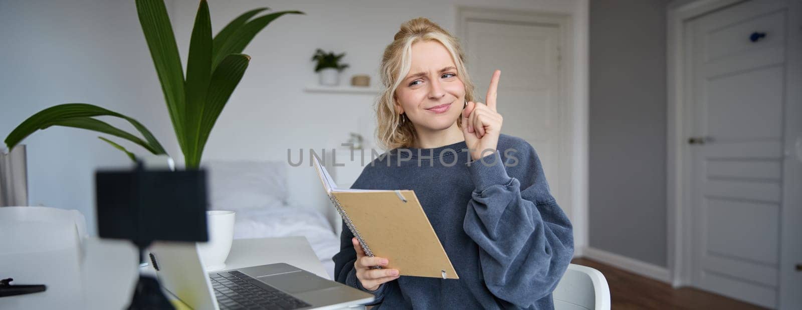 Portrait of young creative woman, content maker, sitting in room, working from home, using laptop, holding notebook, raising finger, has an idea, eureka gesture.