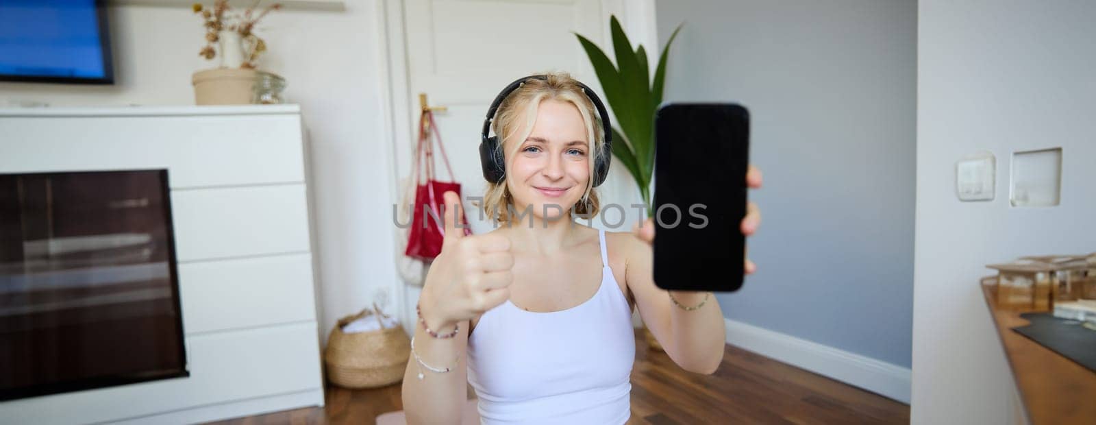 Portrait of young fitness instructor in headphones, showing smartphone screen and thumbs up, recommending app for home workout by Benzoix
