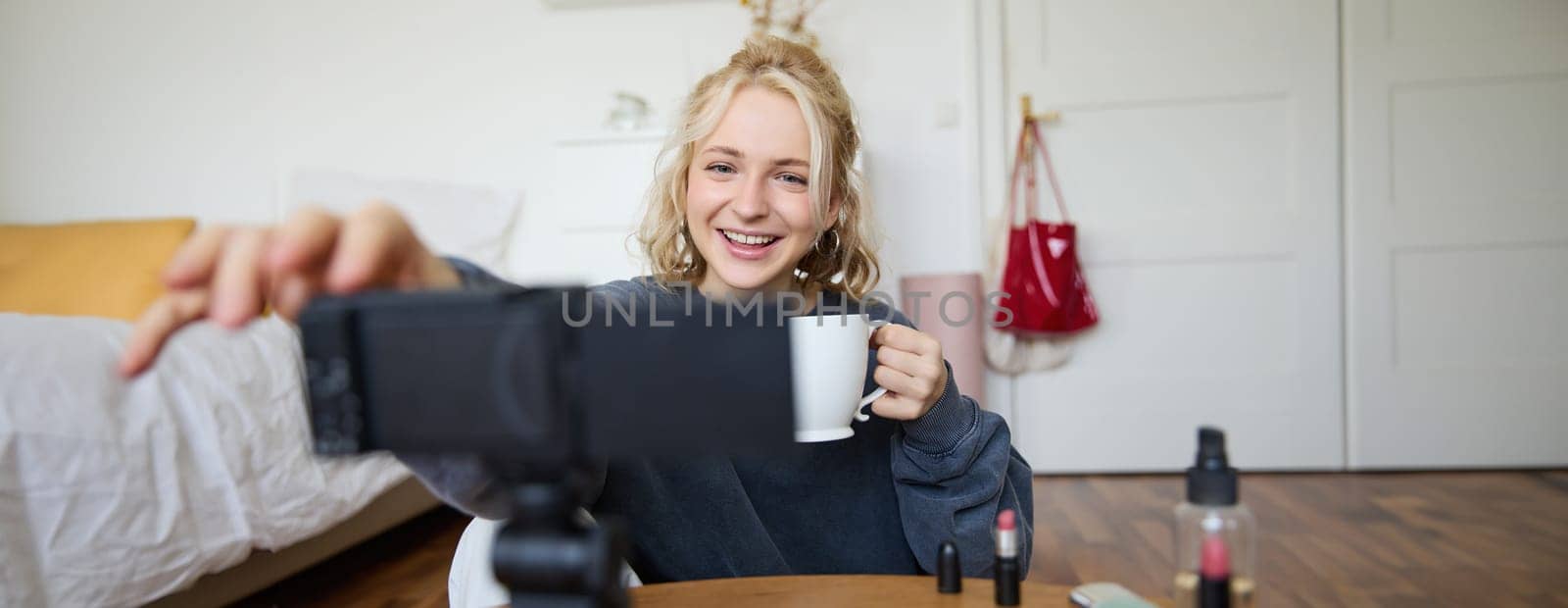 Portrait of cute smiling woman, blond girl drinks tea and records a casual, lifestyle video blog, vlogger sits in a room with camera and stabiliser, holds cup of coffee and talks by Benzoix