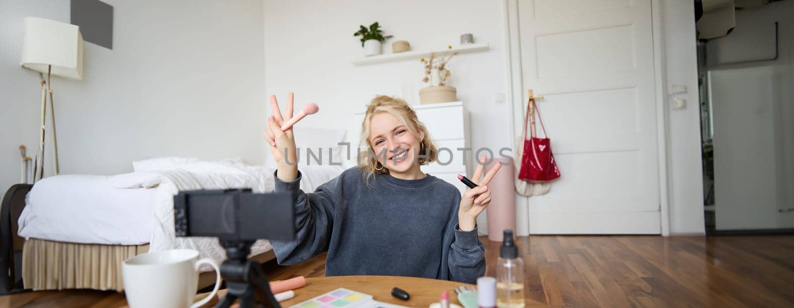 Portrait of young blond woman, teenage girl records video for her social media account, shows makeup on camera, recommends lipstick to online followers, creates content in her room.