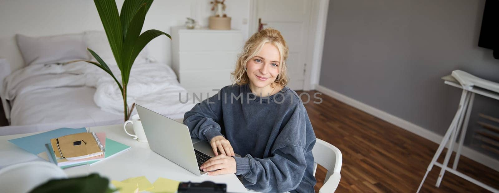 Portrait of young blond woman with laptop, recording video on digital camera, created video blog, vlogging in her room, editing on computer.