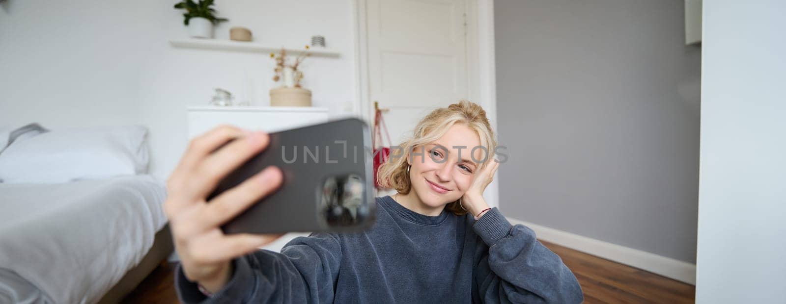 Portrait of young stylish girl sits on bedroom floor, takes selfies on her smartphone, posing for photo on social media app, smiling and looking happy at camera.