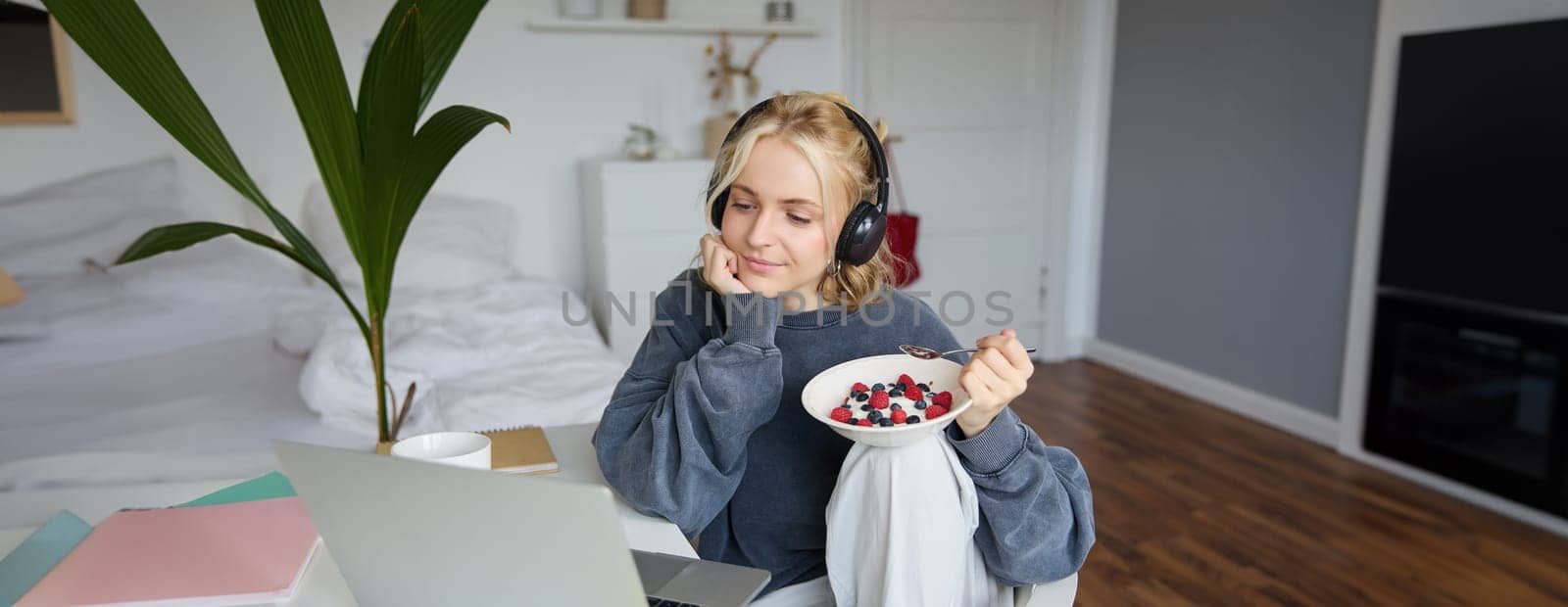 Portrait of happy young blond woman, sitting in a room, watching movie on laptop and eating healthy breakfast, drinking tea, resting on weekend by Benzoix