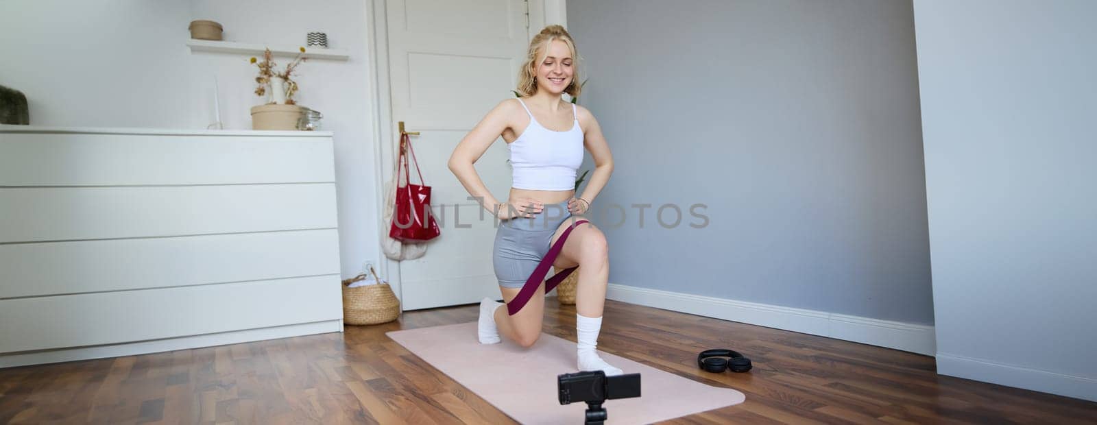 Portrait of young athletic woman recording home workout video, shooting content for sport fitness vlog, using resistance band and digital camera.