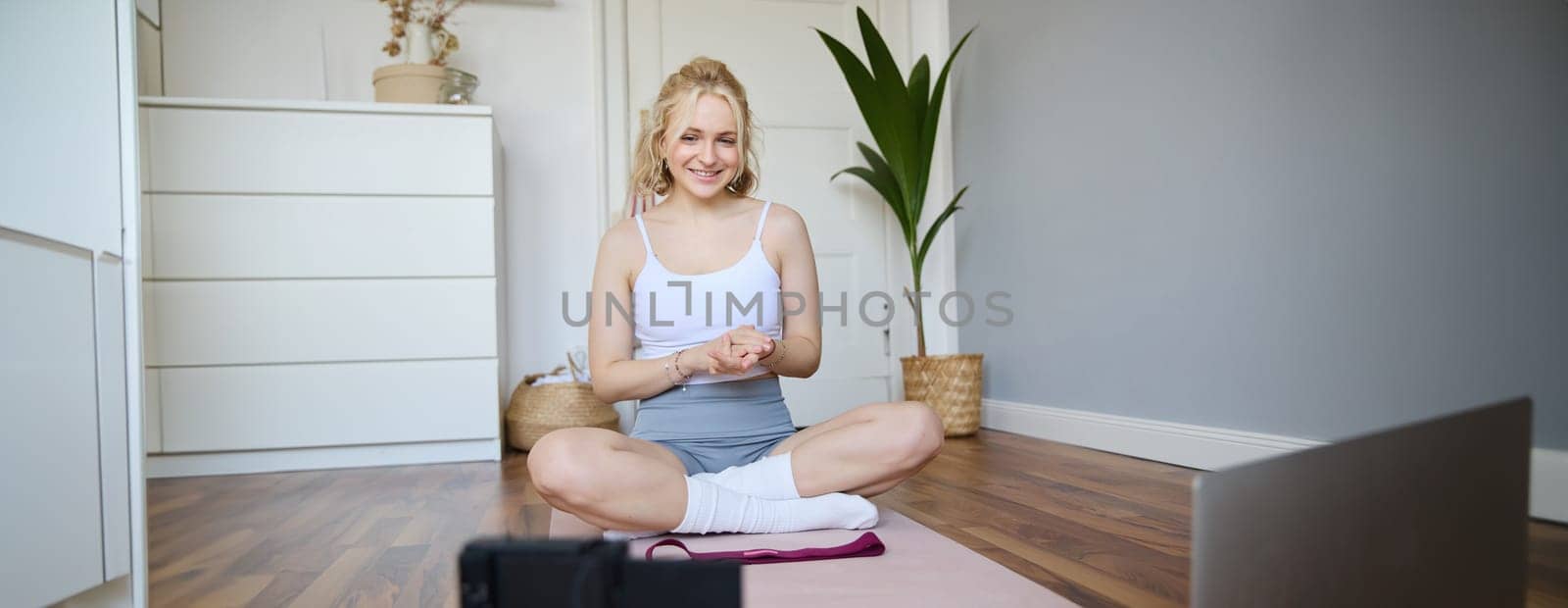 Portrait of young charismatic fitness trainer, girl blogger records video on digital camera, talks about health and workout, doing exercises on rubber mat in a room at home.
