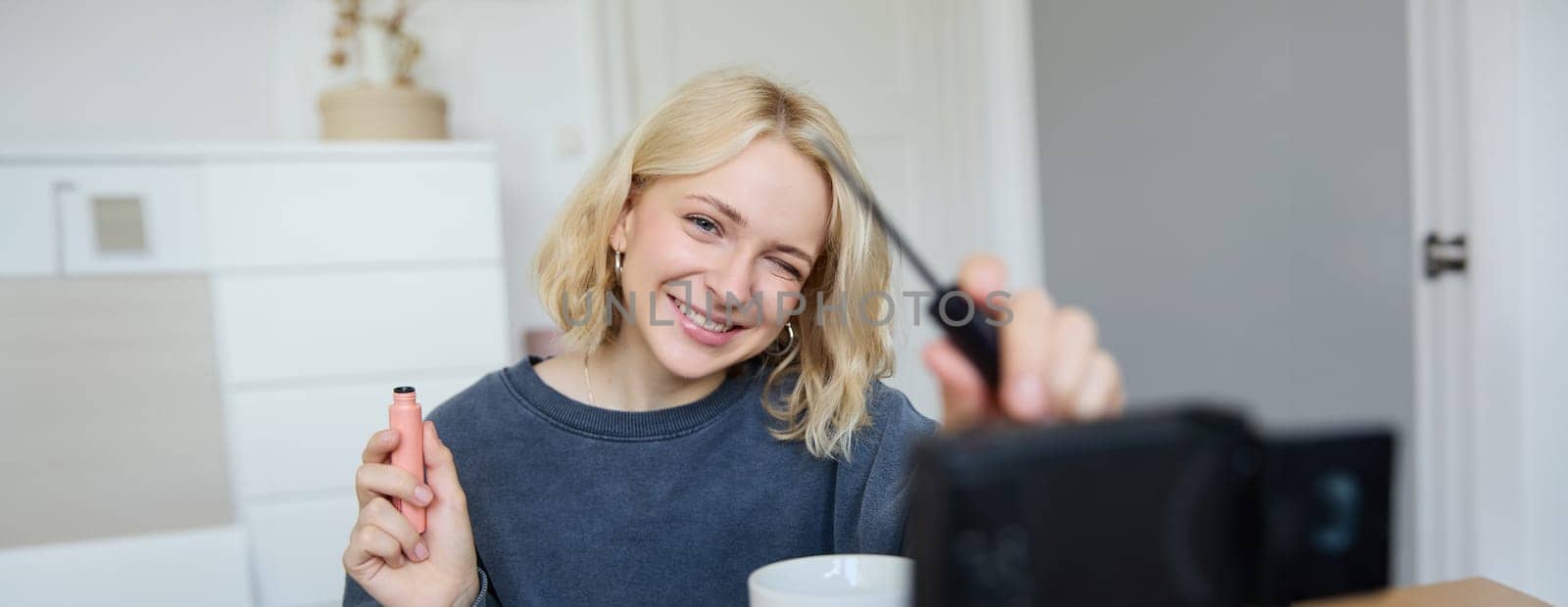 Smiling young beautiful lifestyle blogger, girl records a beauty vlog in her room, using professional video camera, showing makeup cosmetics to her followers, creates content for social media by Benzoix