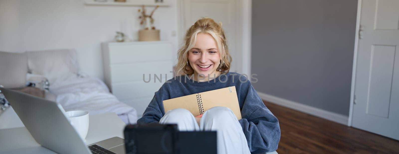 Portrait of blond smiling woman, records video on digital camera how she writes in notebook, talks to followers, doing lifestyle blog content in her room by Benzoix