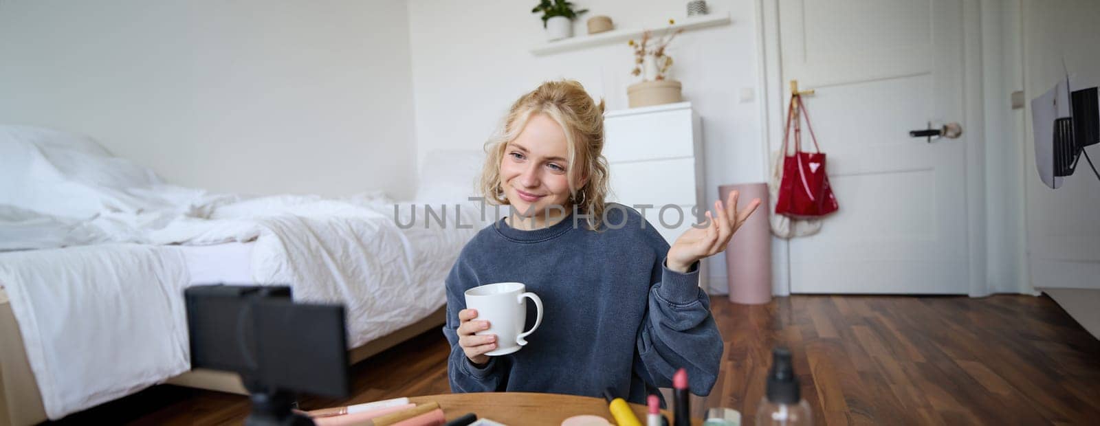 Image of young woman, makeup vlogger, sitting in bedroom with digital camera, drinking tea and talking, creating lifestyle video, social media content by Benzoix