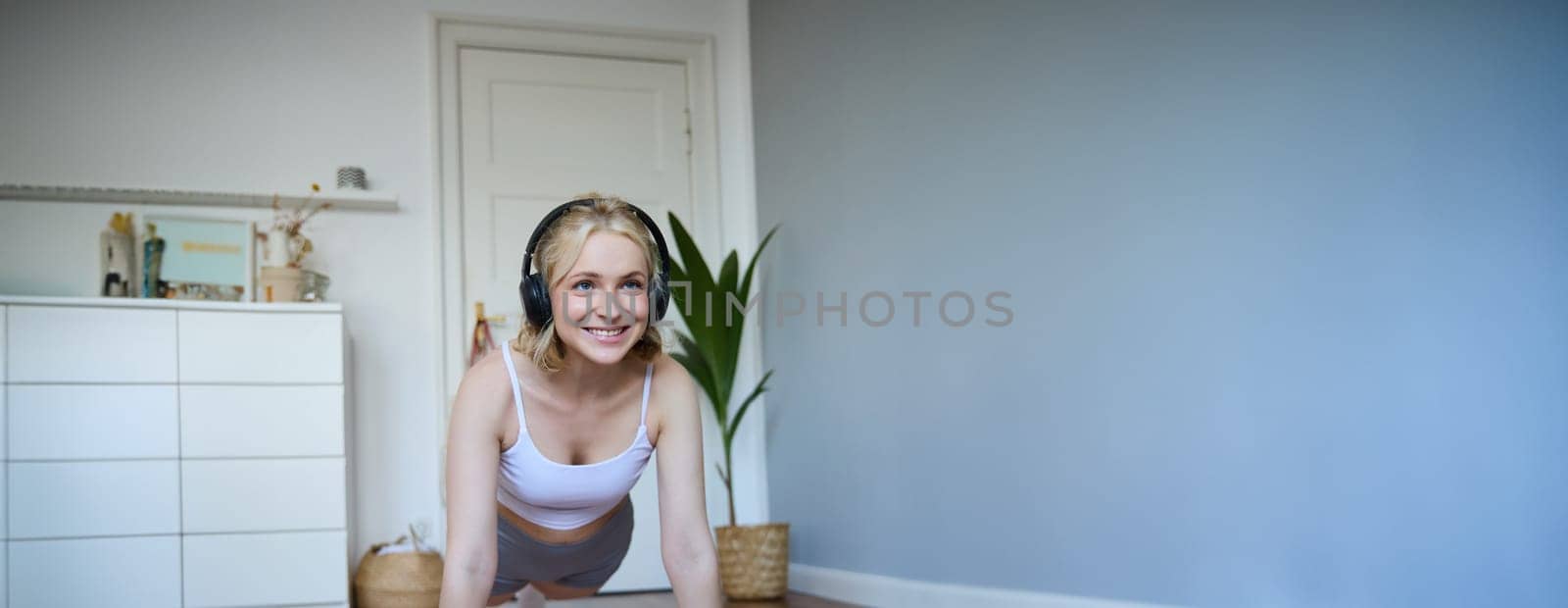 Portrait of young athletic woman in wireless headphones, standing in plank, following online workout video, using laptop to join training session on the internet by Benzoix