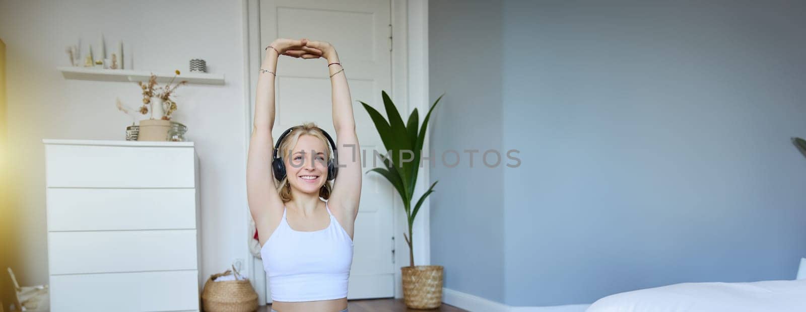 Portrait of woman doing fitness exercises at home, watches sports video on laptop, wearing wireless headphones, follows workout instructions, sits on rubber mat in her room.