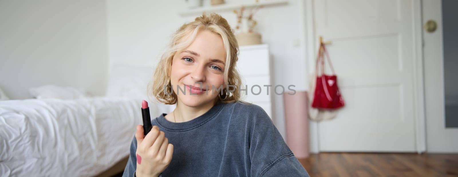 Portrait of young makeup artist, beauty blogger showing new lipstick, recording video in her room, smiling and expressing positivity.