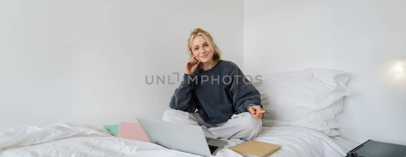Portrait of young woman working from home, sitting on a bed with notebooks, documents and laptop, looking happy and smiling at camera.