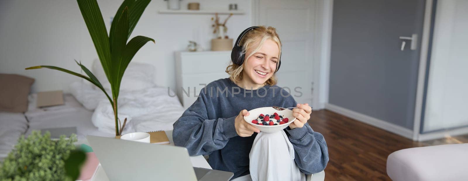 Image of happy woman sitting in a room, watching interesting tv show or movie on laptop, using screaming service, wearing headphones, eating dessert and drinking tea.