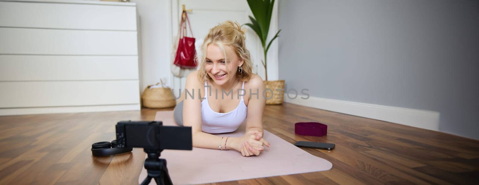Portrait of young fitness instructor, vlogger creating content at home, doing workout and record exercises on digital camera, using rubber yoga mat.
