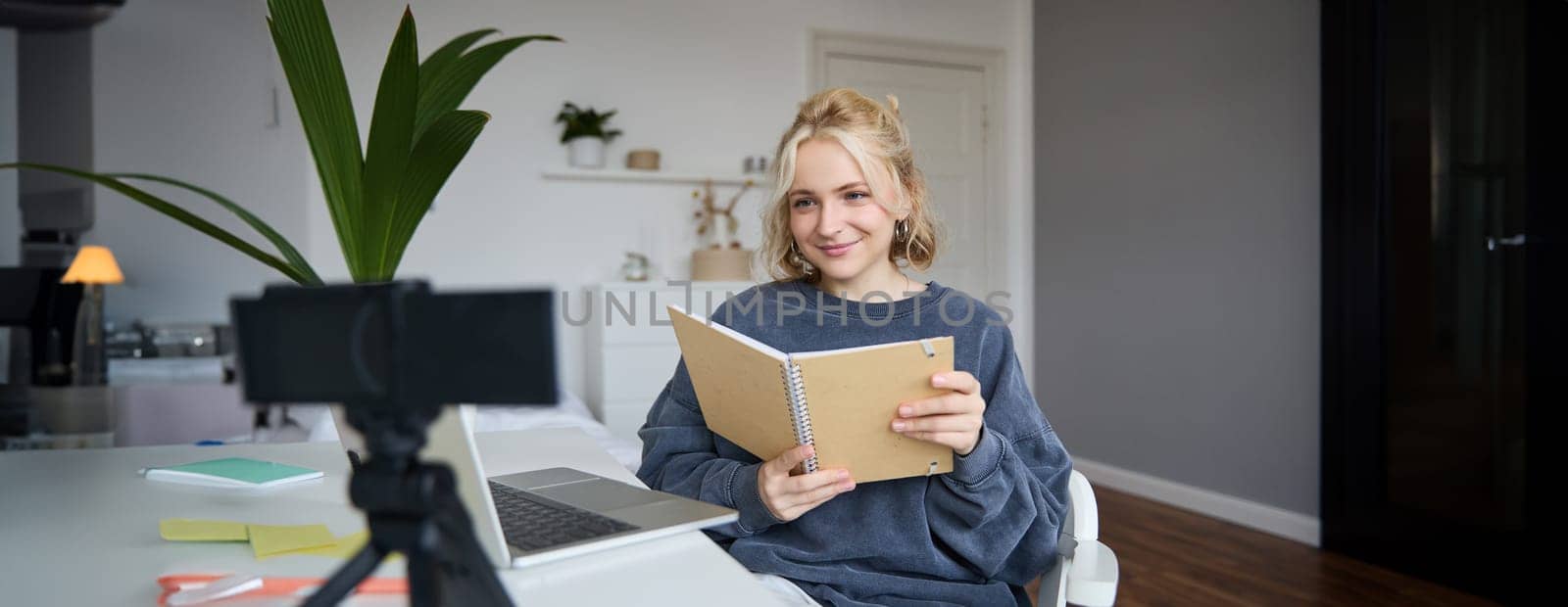 Young smiling blond woman, sits near laptop, uses digital camera to record video blog, creates lifestyle content for social media, records tutorial, shows notebook, reads her notes.