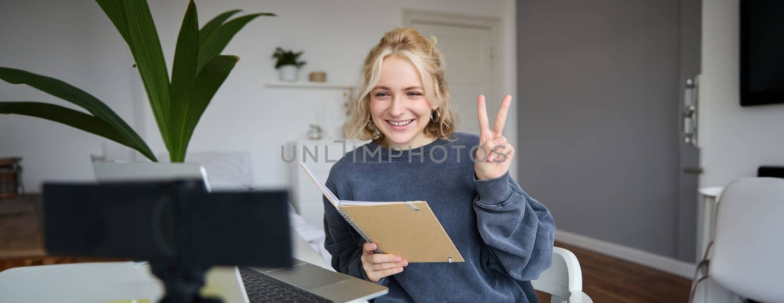 Image of smiling girl records video of herself on digital camera, shows peace sign, sits in front of laptop, holds notebook.