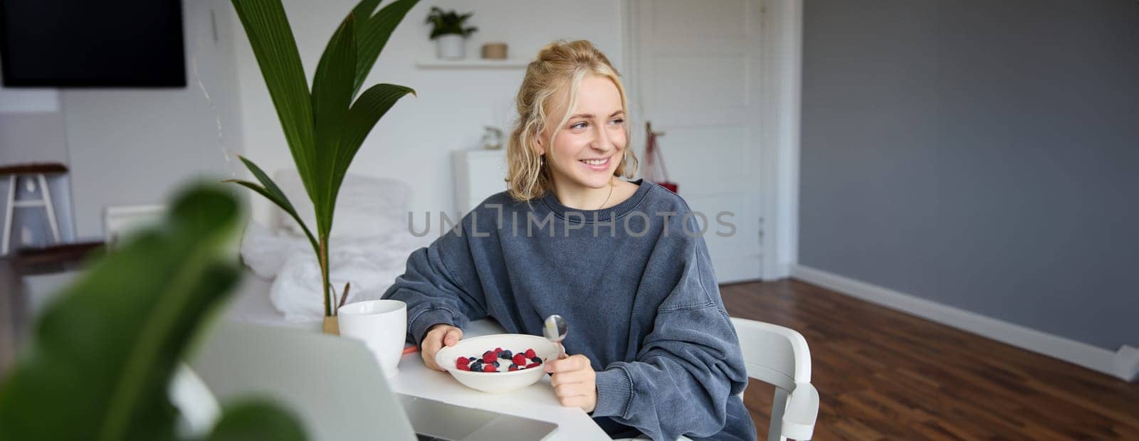 Portrait of young candid girl watching videos on laptop, enjoying movie and eating in front of laptop, having breakfast and staring at screen.