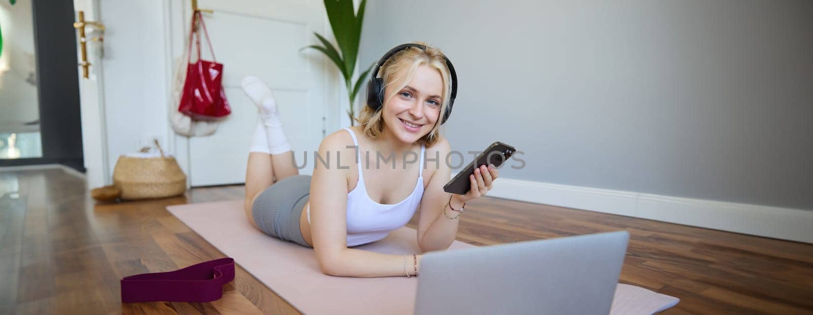 Portrait of young sporty woman, connects to online workout training session, doing exercises, lying on rubber mat, listens to fitness instructor in headphones, holding mobile phone.