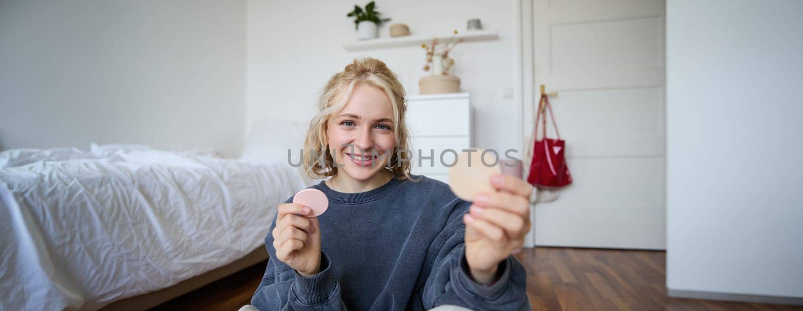 Image of smiling blond woman, vlogger, showing makeup products, beauty items, recommending to followers, sitting on floor in bedroom by Benzoix