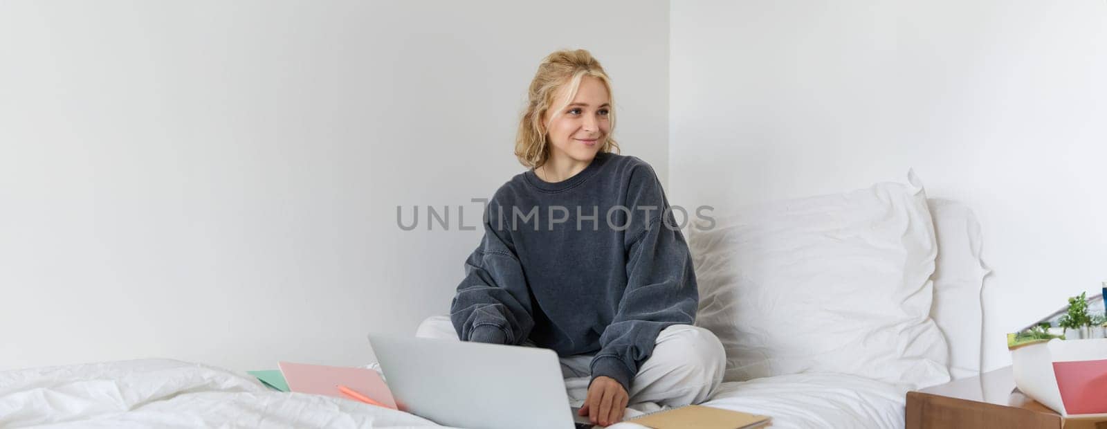 Portrait of happy blond woman, freelancer working from home, sitting on bed with laptop and notebooks. Student doing homework in bedroom, connects to online class via video chat by Benzoix