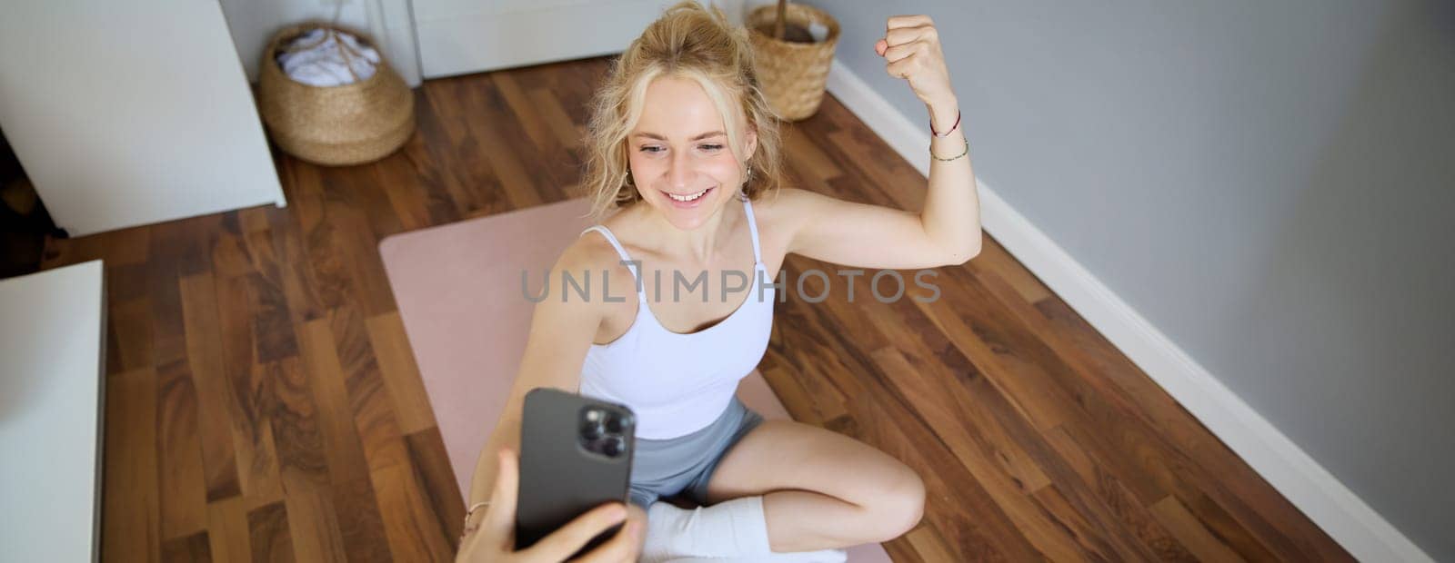 Portrait of young and fit fitness woman, training instructor sitting in a room at home, using yoga mat, flexing biceps, workout, recording herself during exercises by Benzoix