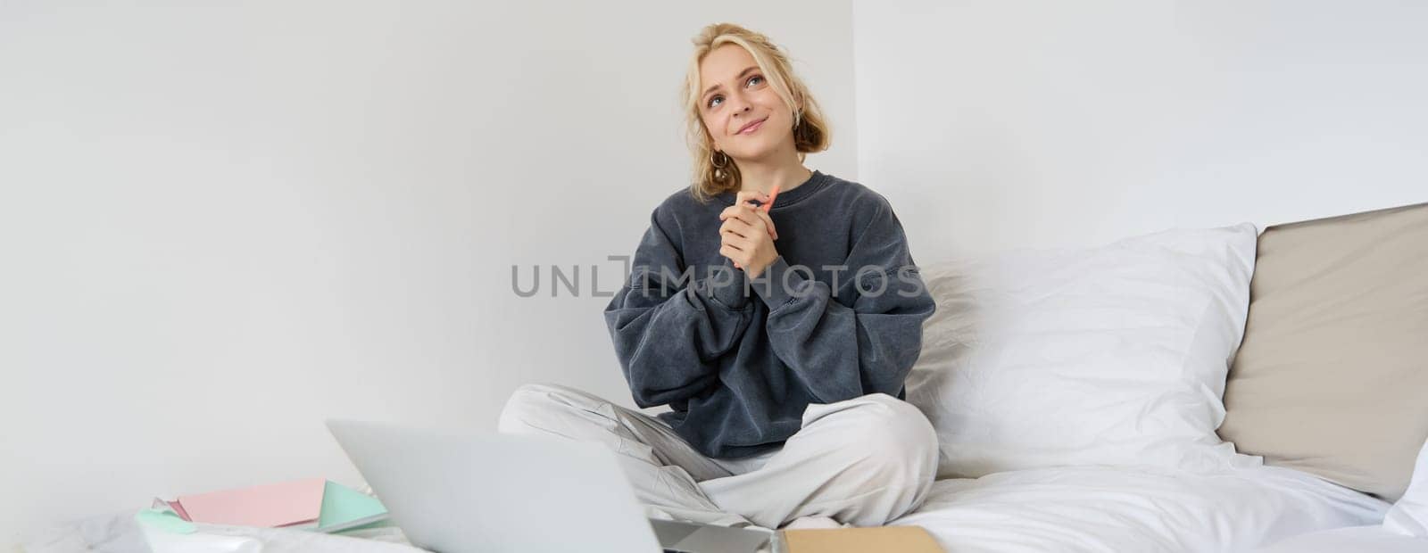 Portrait of cute smiling woman, sitting on a bed, looking dreamy, thinking, using laptop for remote work.