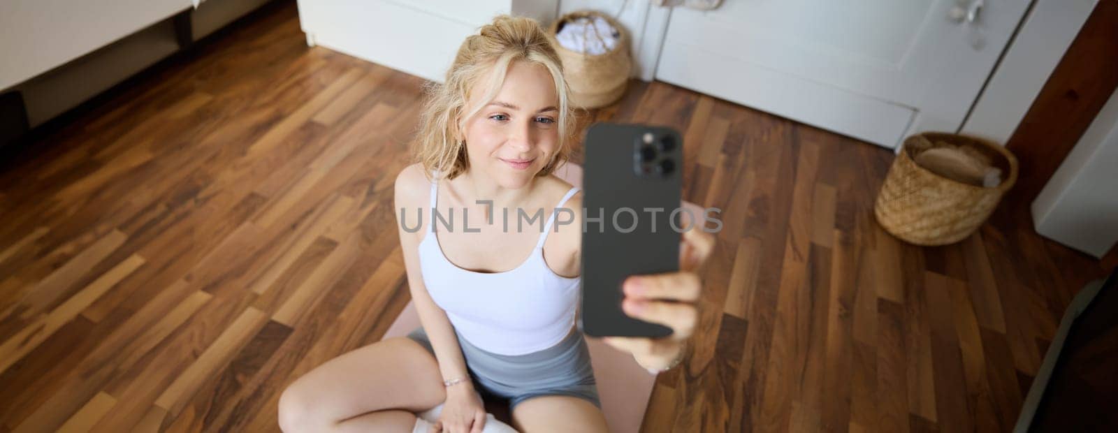 Young sporty fitness trainer, yoga instructor does live stream, takes selfies on smartphone while sitting on rubber mat at home, recording her workout training session by Benzoix