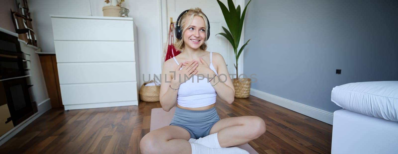 Image of sporty young woman doing workout at home, using rubber yoga mat, listening to music and exercising.