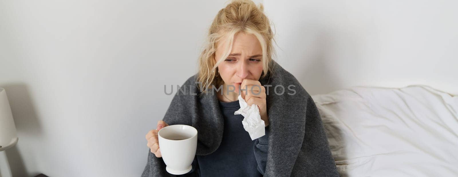 Portrait of woman catching a cold, staying home sick, drinking tea, sneezing in napkin, blowing nose, has covid symptoms by Benzoix