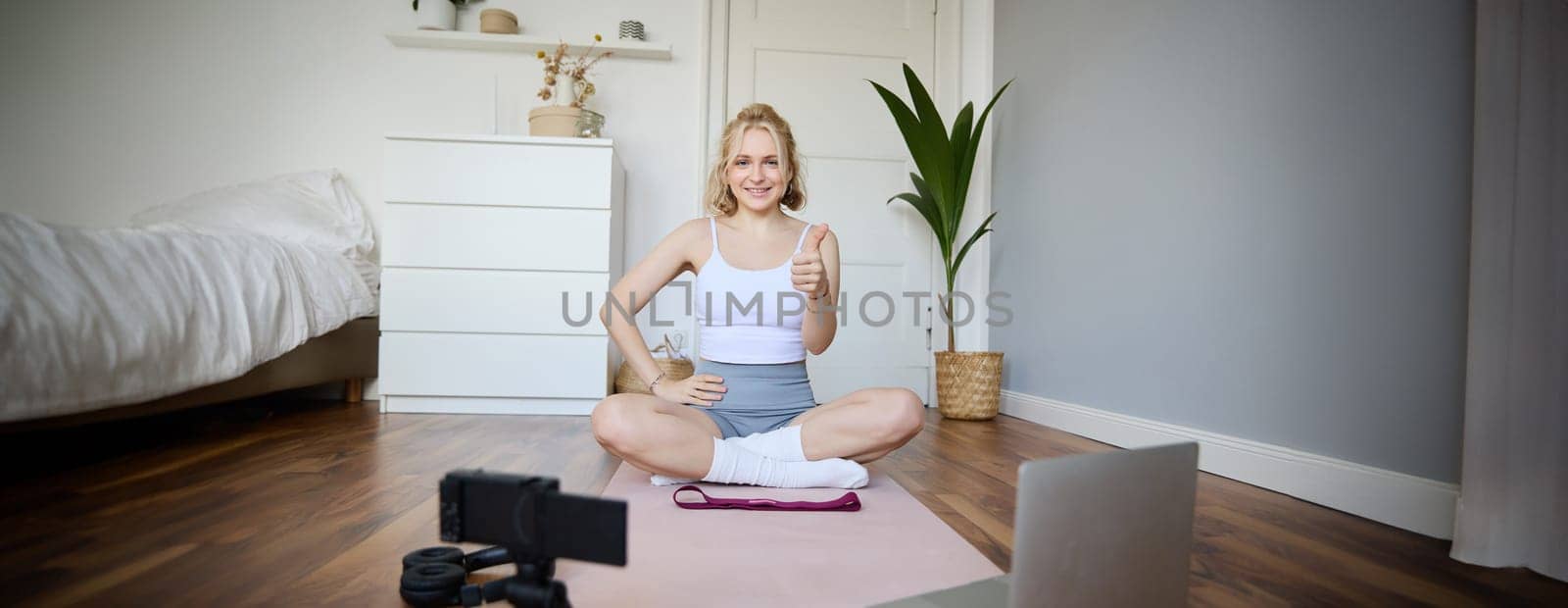 Fitness blogger, woman records video of herself, sitting on rubber mat for exercises in front of digital camera and laptop, shows thumbs up, creates content for social media account about wellbeing.