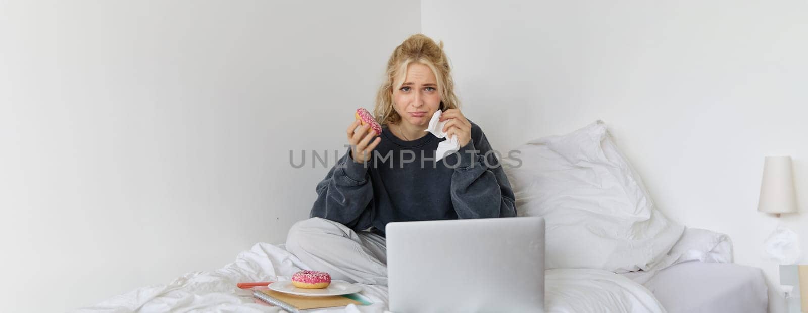 Portrait of sad and depressed woman, feeling heartbroken, lying in bed with comfort food, eating doughnut, crying from upsetting movie, watching something on laptop by Benzoix