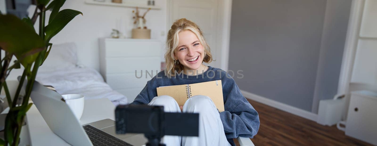 Portrait of cute, smiling young social media content creator, girl records video on digital camera and stabiliser, holds notebook, talks to audience, vlogging in her room.