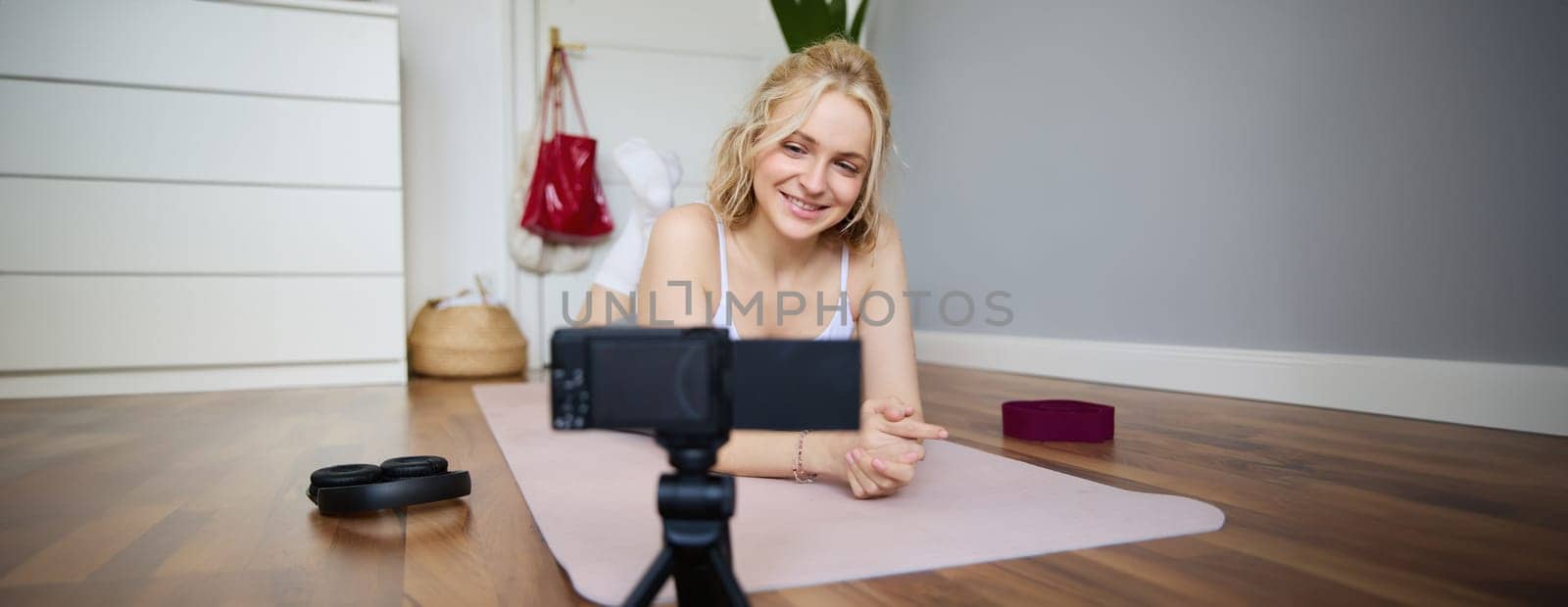 Portrait of young woman, sports vlogger, fitness instructor recording video of herself showing workout exercises, using digital camera, lying on yoga rubber mat by Benzoix