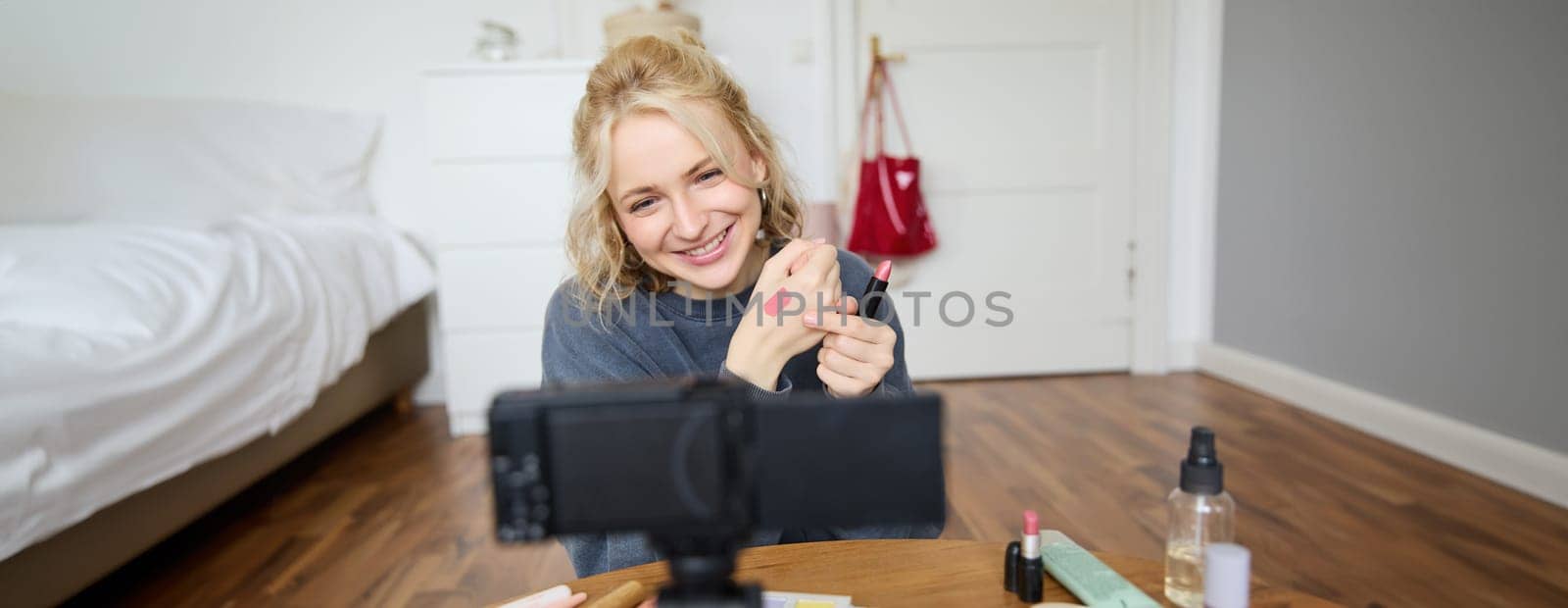 Image of young stylish woman, blogger recording a beauty lifestyle video of her picking best lipstick, showing lip balm swatches on her skin, sitting in front of digital camera in empty room.