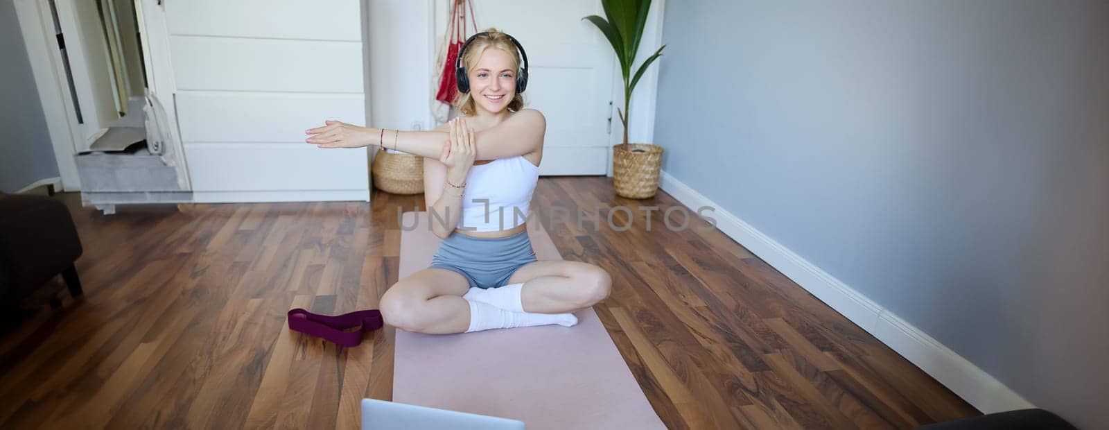 Wellbeing and fitness concept. Woman in headphones, sits on yoga mat, watches workout video on laptop, stretches arms, does exercises.