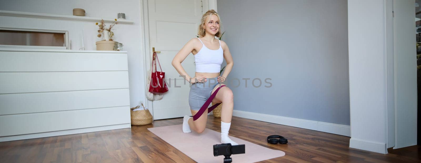 Portrait of fitness instructor, young sporty vlogger woman doing exercises on camera, shooting video about workout, using resistance band and doing sit-ups at home in empty room.