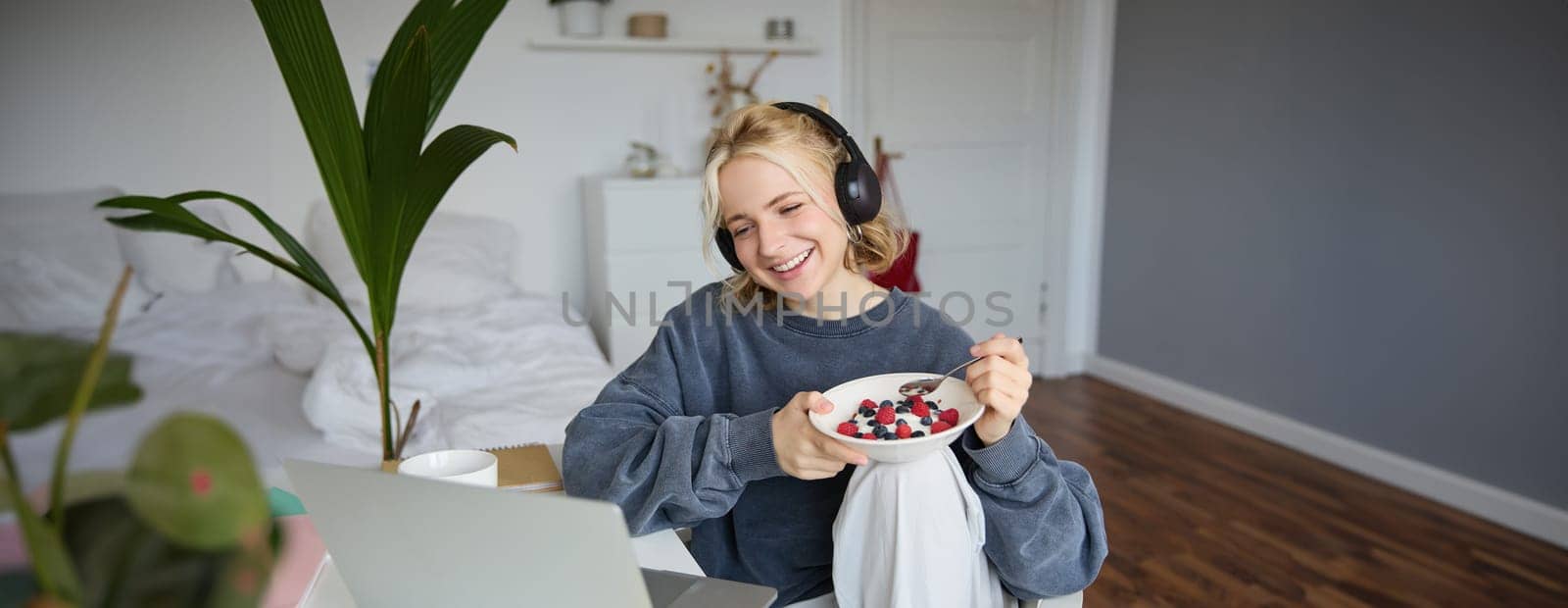 Portrait of smiling beautiful woman, sitting in room with breakfast, eating and watching tv show on laptop, laughing and looking at screen.