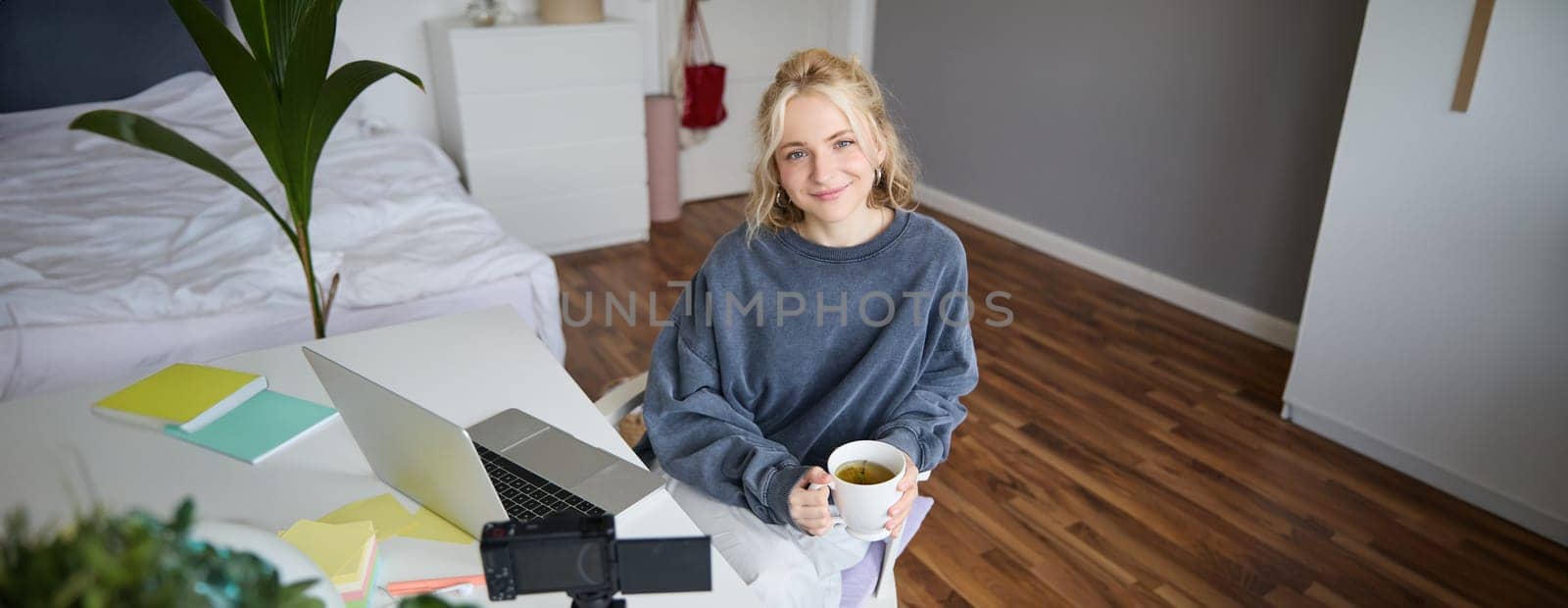 Portrait of young smiling woman, social media influencer, girl with digital camera and laptop, sits with cup of tea in bedroom, records vlog, creates content.