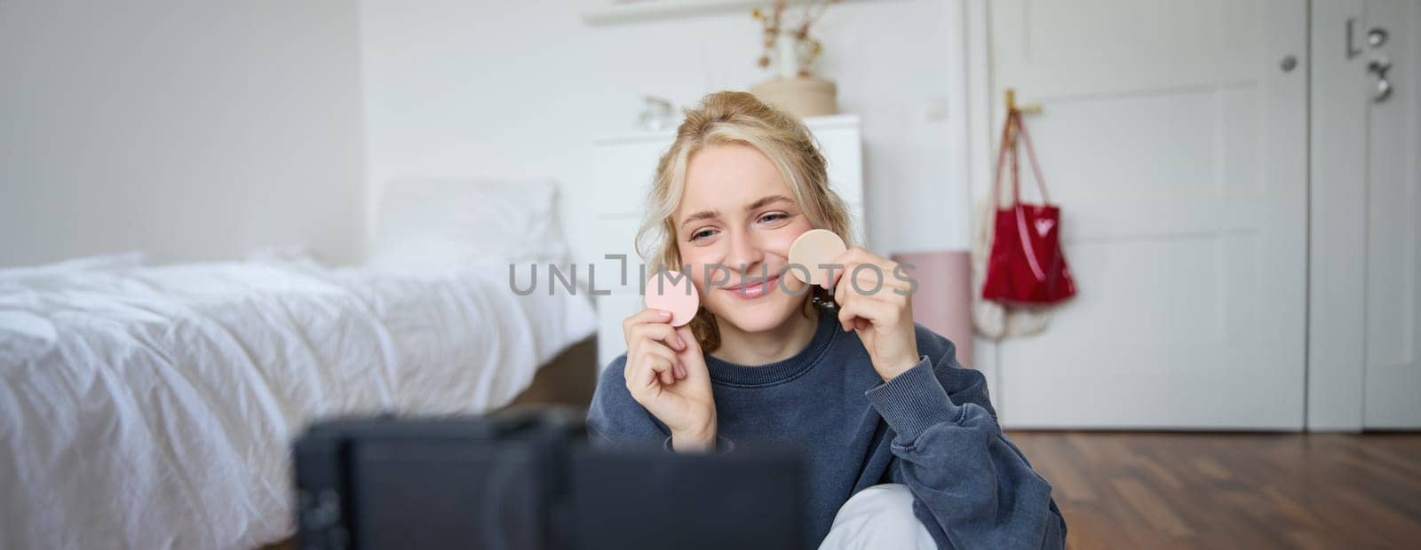 Portrait of young woman, beauty content creator, sitting in a room in front of digital camera, recording makeup tutorial vlog, showing cosmetic facial products.