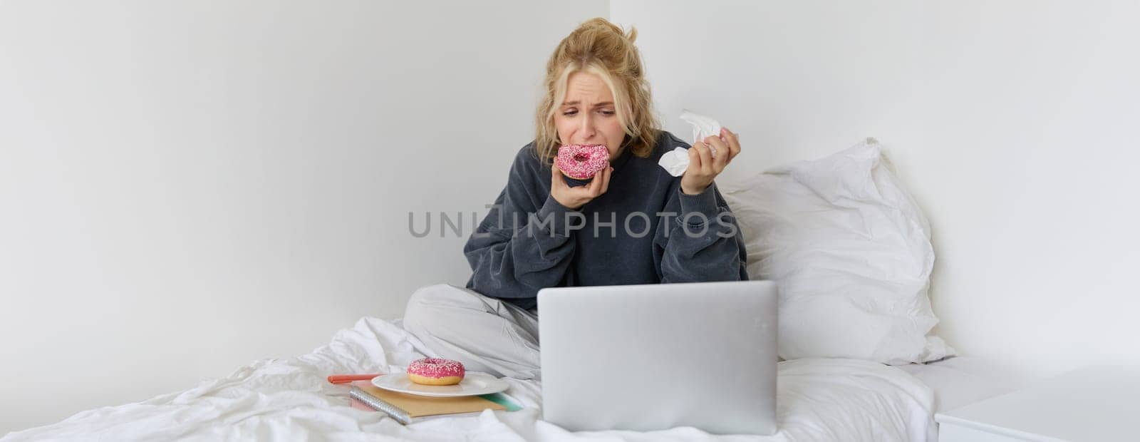 Portrait of sad, crying young woman, staying at home, sitting in bed with doughnut and comfort food, looking at something upsetting on laptop screen.