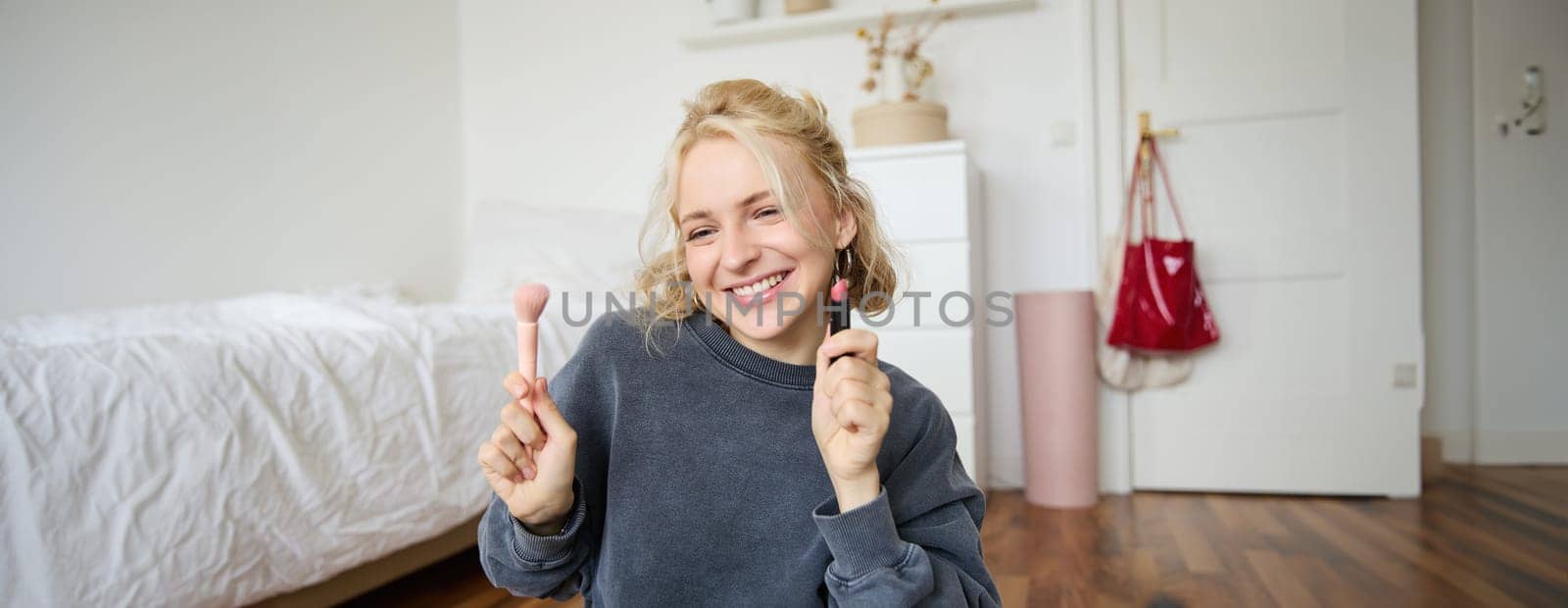 Woman beauty blogger, records video of herself sitting in a room and rating makeup products, puts on make up, holds lipstick and cosmetic brush in hand, using professional camera for content creation by Benzoix