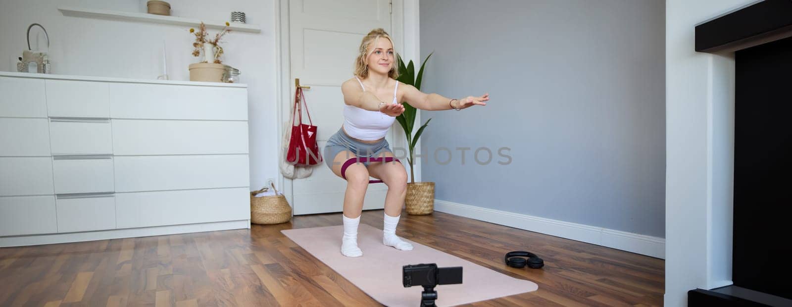 Portrait of woman, fitness instructor at home, recording video about workout, showing how to do leg exercises, squats with elastic resistance band, working out indoors on yoga mat.
