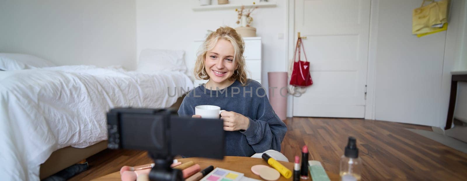 Portrait of young lifestyle blogger, woman sitting in bedroom and talking at digital camera, drinking tea, recording a vlog. Lifestyle content creation