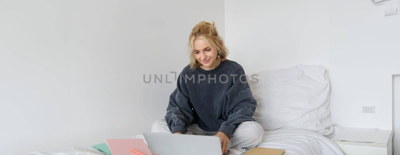 Image of happy young woman, student e-learning from home, connect to online course on her laptop, sits on bed with notebooks, smiling and looking happy by Benzoix
