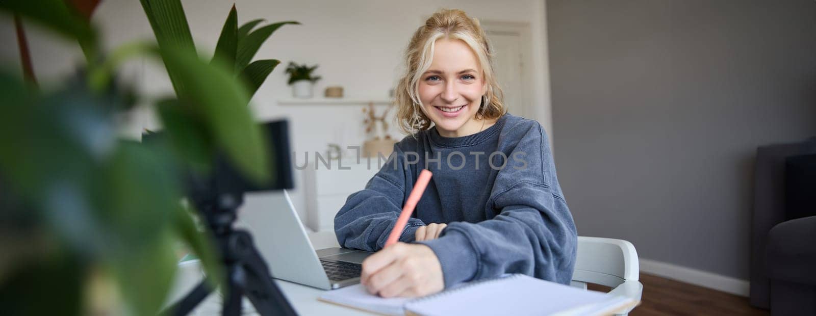 Cute smiling girl sits in a room, writes down notes, doing homework, records video of herself on digital camera, creates content for vlog, lifestyle blogger doing daily routine episode.