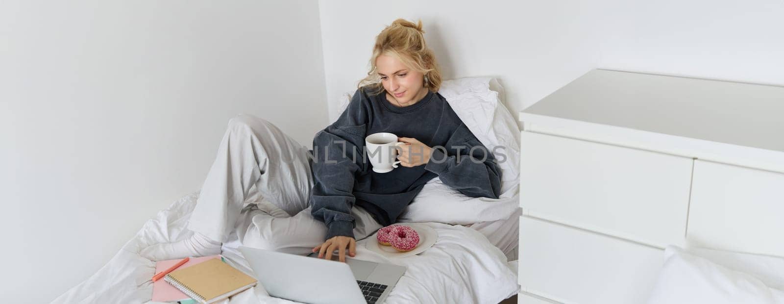 Portrait of young beautiful blond woman, feeling lazy at home, lying in bed with cup of tea, doughnut and laptop, studying remotely or watching movie online.